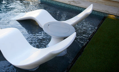 Two sculptural white chaise lounges and a small round table positioned in a shallow pool beside a grassy edge