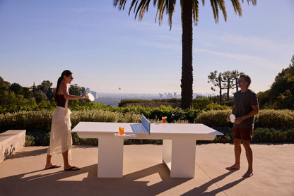 Two people playing ping pong on a white outdoor table with a city view and palm tree backdrop.