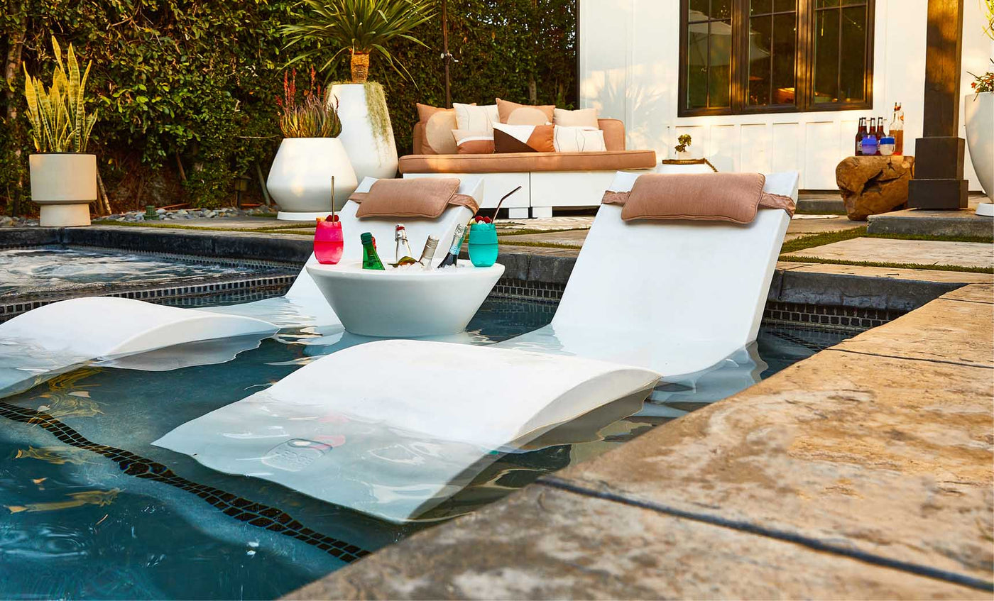 White poolside ice bin table filled with drinks, partially submerged in shallow water beside a lounge chair.