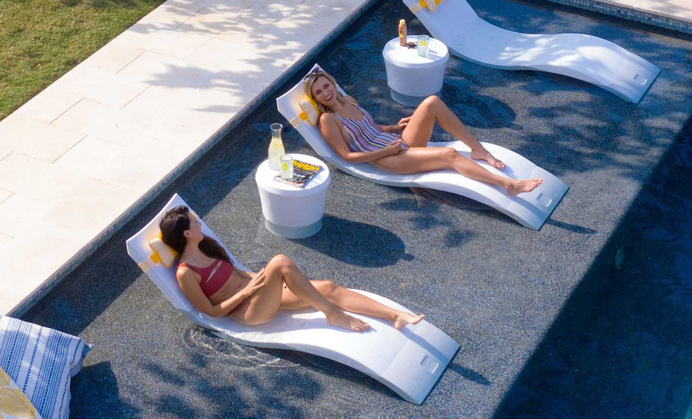 White chaise loungers in a shallow sun shelf pool with women relaxing beside small side tables.