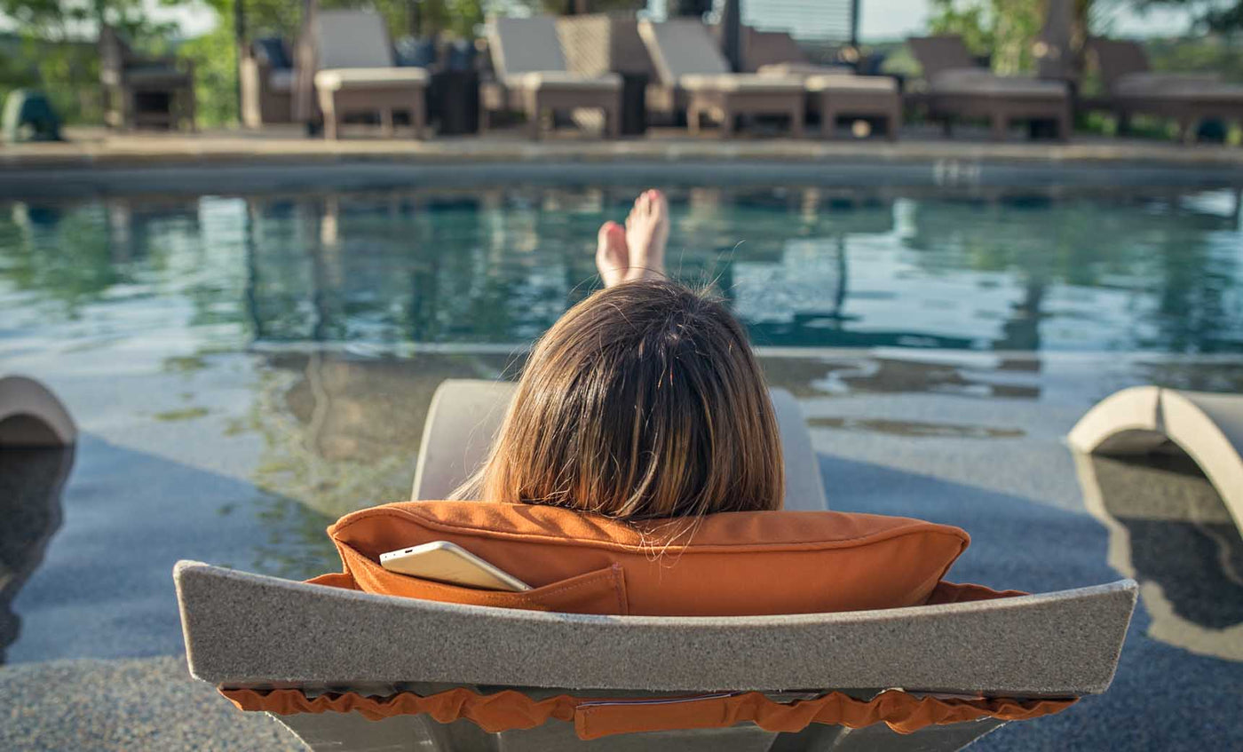 Person reclining on an in-pool lounge chair with orange cushion and phone pocket by the pool.