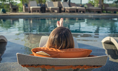 Person reclining on an in-pool lounge chair with orange cushion and phone pocket by the pool.