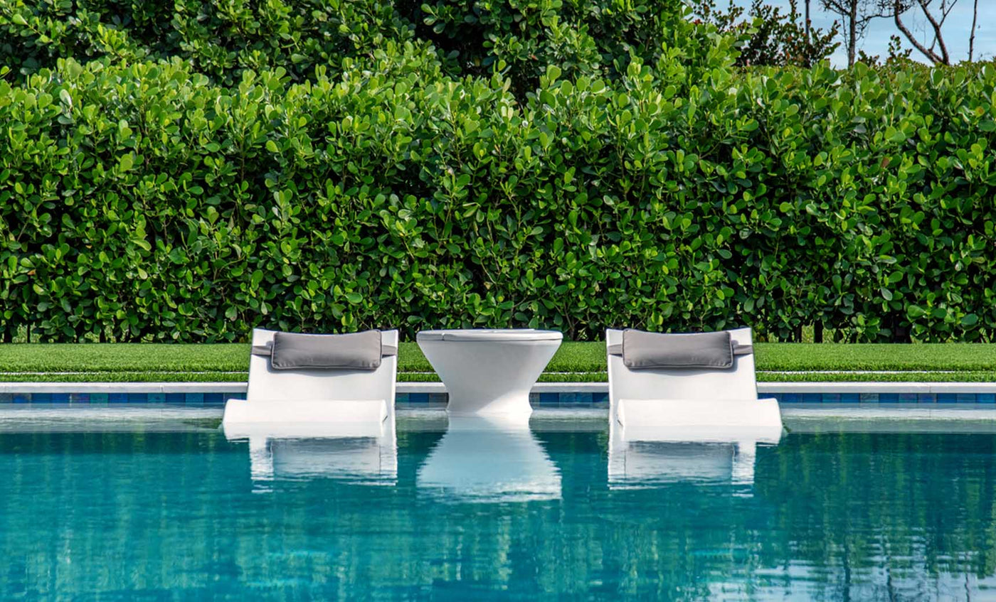 Two white in-pool lounge chairs with gray headrests and a white side table in shallow pool water.