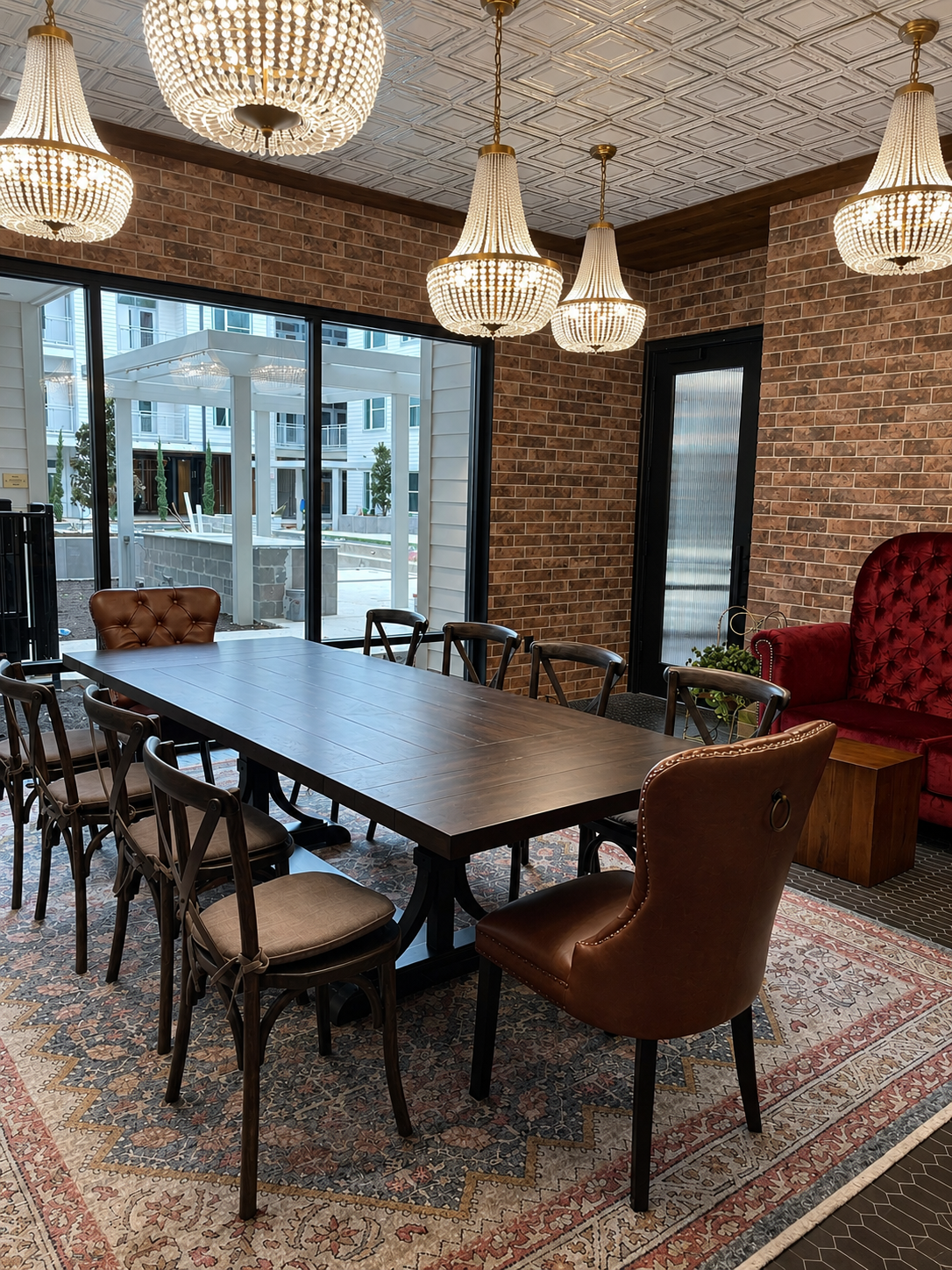 Dining room with a long wooden table and chairs, featuring chandelier lights and a brick wall.